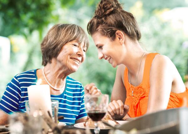 A mother and her adult daughter enjoy a meal and a glass of wine outdoors at a restaurant.