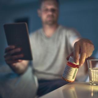 : A man sits up in bed to check a medication on his side table.