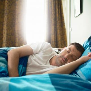 A man sleeps peacefully on his side while the morning sun peeks through his bedroom curtains.