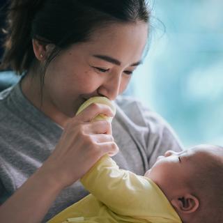 A smiling mother holds her young baby and kisses the baby’s hand.