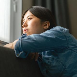 A young woman inside her home looks out her window longingly.